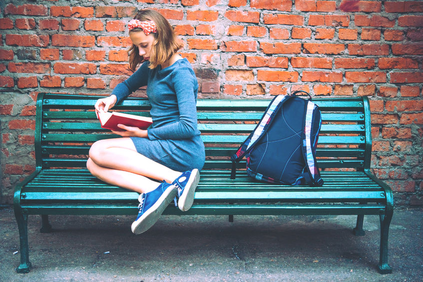 50840812 - a teenage girl is reading on a bench with brick wall in the background. toned image